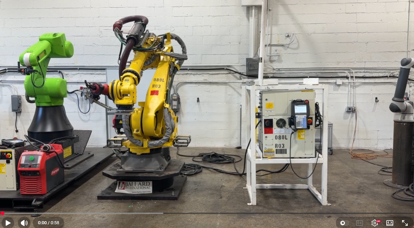 Industrial robotics lab with a yellow robotic arm and a green robot arm on a workbench area, cables on the floor