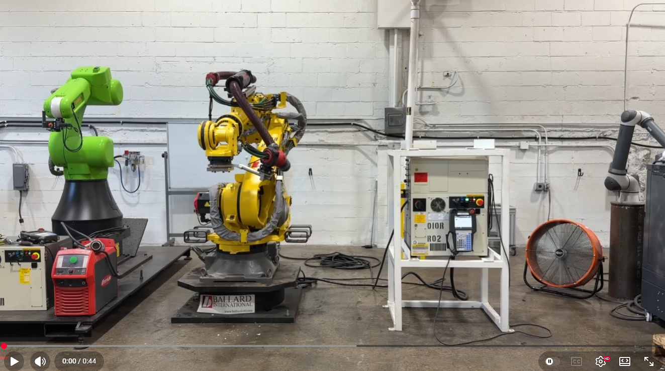 Industrial robotics lab with a bright green robotic arm on the left and a yellow robotic arm center-right on a platform, connected to cables and control units against a white brick wall.